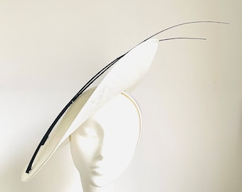 May include: A wide-brimmed white hat with a smooth, structured surface and a thin, black curved accent piece extending from the top. The hat is mounted on a plain white display head, showcasing its angled, asymmetrical silhouette and clean lines.