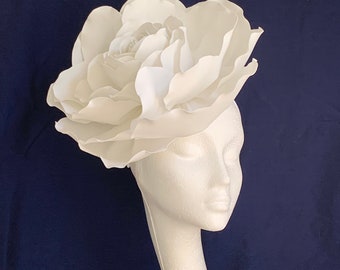 May include: A large white fabric flower, resembling a rose, is worn as a hat on a white mannequin head.