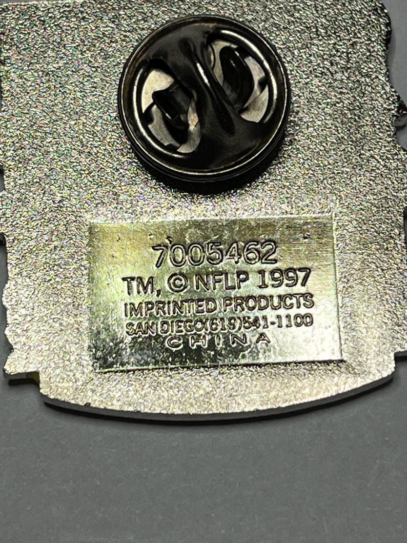 May include: A silver-toned metal pin with a black clasp. The pin has a textured background and a rectangular section with the text "7005462 TM, &copy; NFLP 1997 IMPRINTED PRODUCTS SAN DIEGO(619)541-1100 CHINA".