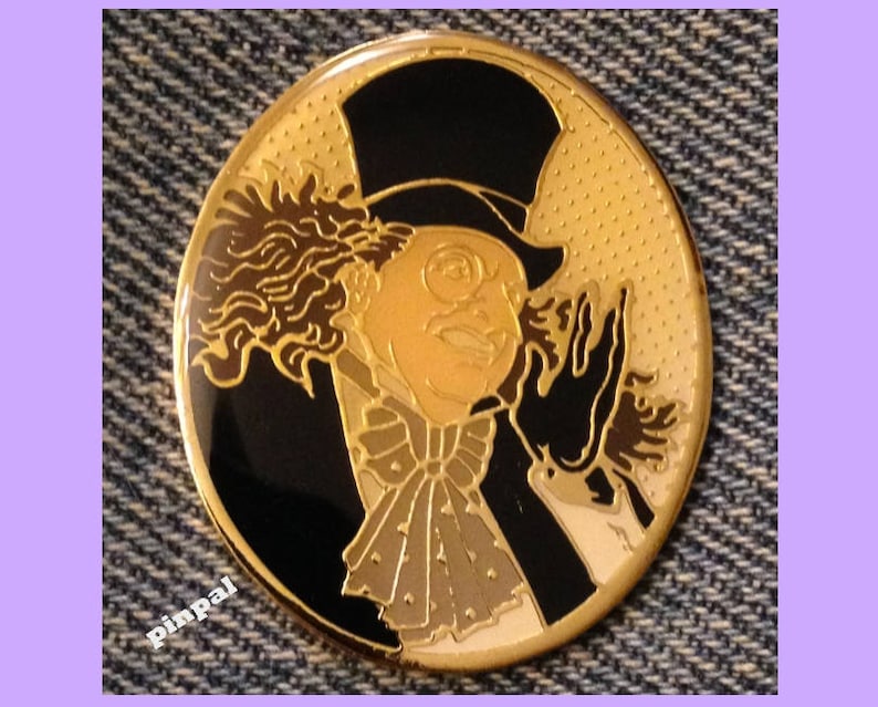 May include: A gold-colored metal pin featuring a cartoon image of the Penguin from Batman. The Penguin is wearing a black top hat, a black tuxedo, and a white bow tie. He has a white face with black eyes and a black mustache. The background is a black circle with a gold-colored dotted pattern.