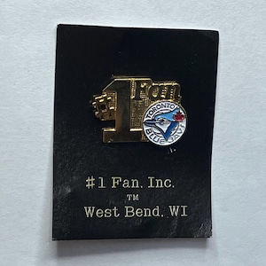 Può includere: Spilla dorata con il numero 1 e la scritta "#1 Fan" e il logo dei Toronto Blue Jays. La spilla è su una carta nera con la scritta "#1 Fan, Inc. West Bend, WI."