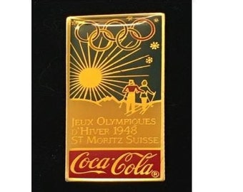 May include: A gold-colored pin with a depiction of the 1948 Winter Olympics in St. Moritz, Switzerland. The pin features the Olympic rings, a sun, two skiers, and the Coca-Cola logo.