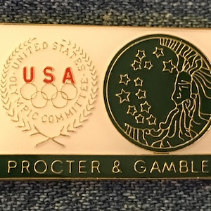 Puede incluir: Un pin de color dorado con un fondo blanco que presenta el logotipo del Comité Olímpico de los Estados Unidos y el texto "USA". El lado derecho del pin presenta un círculo verde con una luna creciente y estrellas, y el texto "PROCTER & GAMBLE" está impreso debajo.
