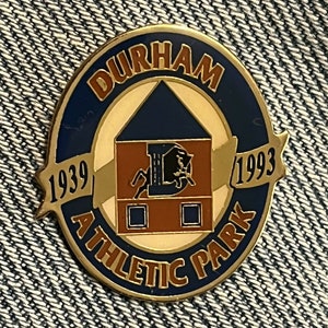 May include: A gold-coloured pin with a blue background and the text "Durham Athletic Park" in a gold circle. The centre of the pin features a brown building with a white roof and a black bull. The years "1939" and "1993" are on either side of the building.
