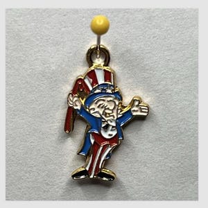 Puede incluir: Encanto dorado con un dibujo animado del Tío Sam con un sombrero y un traje rojo, blanco y azul. Lleva un palo en la mano izquierda y saluda con la mano derecha.