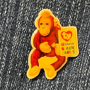 May include: A colorful orangutan-shaped pin with a yellow border. The orangutan is wearing a red outfit and holding a yellow tag that reads "ty teenie BEANIE BABIES". The pin is set against a dark blue denim background.