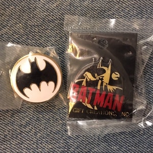 Op de afbeelding: Twee Batman-pins. E&eacute;n pin heeft een zwart-wit Batman-logo op een witte achtergrond. De andere pin heeft een gele Batman met een rode "BATMAN"-tekst en de tekst "GIFT CREATIONS, INC." eronder.