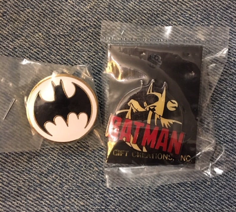 Op de afbeelding: Twee Batman-pins. E&eacute;n pin heeft een zwart-wit Batman-logo op een witte achtergrond. De andere pin heeft een gele Batman met een rode "BATMAN"-tekst en de tekst "GIFT CREATIONS, INC." eronder.
