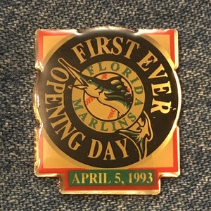 Peut inclure: Épinglette dorée avec un cercle noir représentant un marlin et le texte "FIRST EVER OPENING DAY" avec les mots "FLORIDA MARLINS" en cercle autour du poisson. Le texte "APRIL 5, 1993" est sur une bannière verte en bas.
