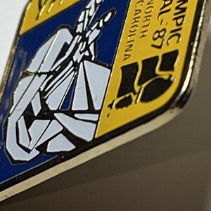 May include: A yellow and blue enamel pin with a black silhouette of a person running. The text "Olympic Trial - 87" and "North Carolina" are on the pin.