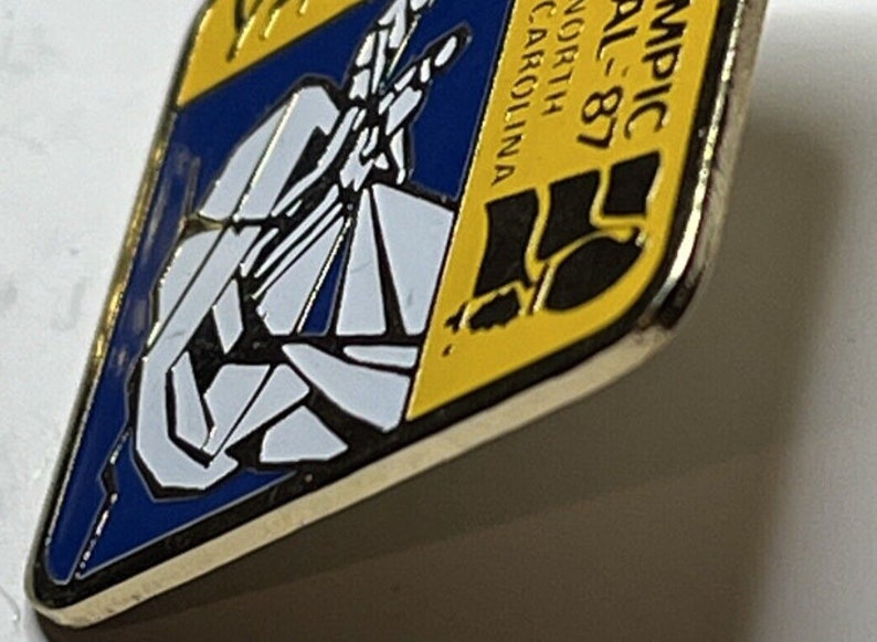 May include: A yellow and blue enamel pin with a black silhouette of a person running. The text "Olympic Trial - 87" and "North Carolina" are on the pin.