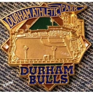 May include: A gold-coloured pin featuring a blue and white banner that reads "Durham Athletic Park". The pin depicts a baseball field with a green outfield and a brown infield. The bottom of the pin reads "Durham Bulls" in blue letters with a red outline.