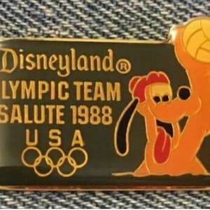 May include: A rectangular pin with a gold border and a dark green background. The pin features the text "Disneyland Olympic Team Salute 1988 USA" in gold lettering, along with the Olympic rings and a cartoon image of Pluto holding a volleyball.