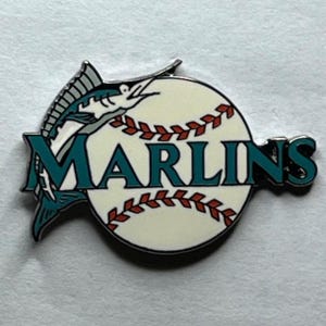 May include: A teal blue and white baseball pin featuring a marlin fish and the word "MARLINS" in teal blue letters.
