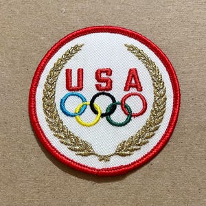 May include: Embroidered patch featuring the Olympic rings and the word "USA" in red capital letters. The patch has a white background, a red border, and a gold laurel wreath design. The Olympic rings are in blue, yellow, black, green, and red.