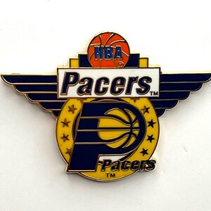 May include: A gold-colored pin featuring the Indiana Pacers logo. The pin has a yellow background with a blue 'P' and basketball design. The word "Pacers" is written in white, with "NBA" and a basketball at the top.