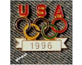 May include: A vintage pin featuring the Olympic rings and the text "USA" in red letters. Below the rings is a white banner with the year "1996" in black. The pin has a gold border and is set against a textured background.
