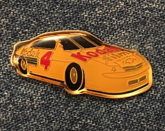 May include: A gold-colored pin of a yellow race car with the number 4 on the side. The car has a black windshield and black tires. The car has the words "Kodak film" on the side in red and black letters.