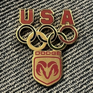 Peut inclure: Une épingle dorée avec le mot "USA" en lettres rouges, des anneaux olympiques et le mot "DODGE" en lettres rouges avec un logo de tête de bélier doré.