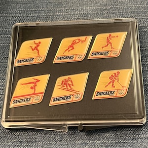 Könnte beinhalten: Ein transparentes Plastiketui mit sechs goldenen Anstecknadeln, die verschiedene olympische Sportarten zeigen. Jede Nadel trägt das Snickers-Logo sowie die Aufschriften "USA" und "Brand".