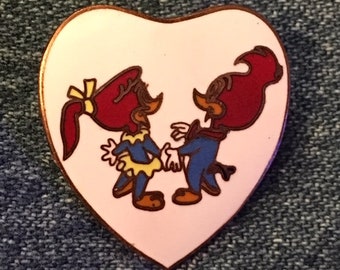 May include: A heart-shaped pin featuring Woody Woodpecker and his girlfriend, Winnie. The cartoon characters are depicted holding hands against a light pink background, with a copper-colored border. The pin is set against a denim background.