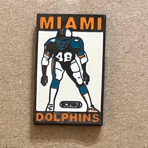 May include: A rectangular pin featuring a Miami Dolphins football player in a uniform with the number 48. The pin has the words "MIAMI" and "DOLPHINS" in orange lettering. The player is in a stance, ready to play.