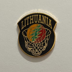 May include: A black and gold Lithuania basketball pin. The pin features the word "LITHUANIA" in gold lettering above a basketball net with a colorful design inside. The design includes green, yellow, and red elements.