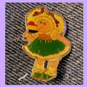 Può includere: Uno spillo dorato con una ragazza dei cartoni animati con i capelli biondi, che indossa un vestito verde e tiene un lecca-lecca con un design rosso, giallo e nero.