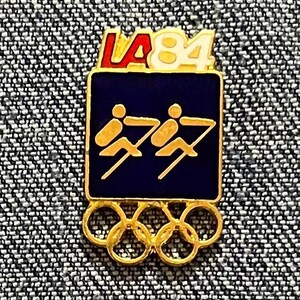May include: A vintage pin featuring the LA 1984 Olympic Games logo. The pin has a rectangular shape with a dark blue background and gold figures of rowers. The Olympic rings are at the bottom, and the text "LA 84" is at the top.