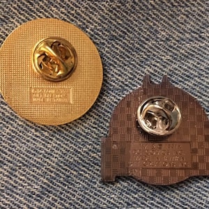 Op de afbeelding: Twee gouden metalen pinbadges. De eerste badge is rond en heeft een glad, getextureerd oppervlak. De tweede badge is gevormd als een gebouw en heeft een getextureerd oppervlak met een geruit patroon. De achterkant van de tweede badge heeft de tekst "IV & 191K, DC Comics Inc., 1992 TM & &copy; 1992, DC Comics Inc., Made in Taiwan".