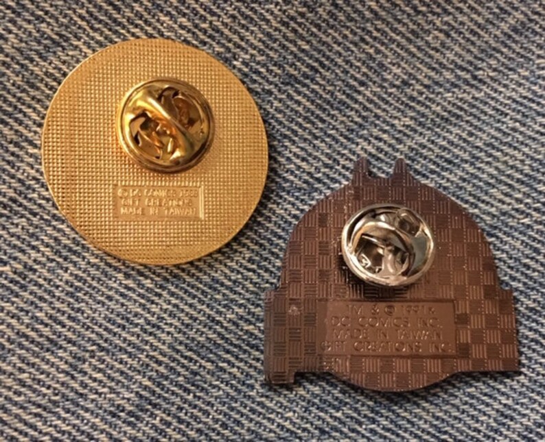 Op de afbeelding: Twee gouden metalen pinbadges. De eerste badge is rond en heeft een glad, getextureerd oppervlak. De tweede badge is gevormd als een gebouw en heeft een getextureerd oppervlak met een geruit patroon. De achterkant van de tweede badge heeft de tekst "IV & 191K, DC Comics Inc., 1992 TM & &copy; 1992, DC Comics Inc., Made in Taiwan".