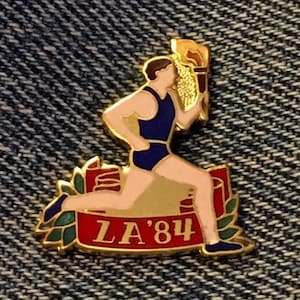 May include: Gold-toned pin featuring a runner holding a torch, with the text "LA'84" on a red and gold banner.