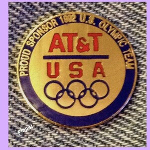 May include: A gold-colored pin with a blue and white border. The pin features the text "Proud Sponsor 1982 U.S. Olympic Team" and the logo "AT&T USA" with the Olympic rings below.