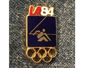 May include: A gold-colored pin with a blue background and a white and red "LA84" inscription. The pin depicts a stylized figure rowing a boat. The pin also features five interlocking gold rings, representing the Olympic Games.