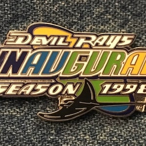 Peut inclure: Épinglette métallique avec l'inscription "Devil Rays Inaugural Season 1998". L'épinglette présente un motif de raie noire et est dans les tons de bleu, vert, jaune et blanc. L'épinglette est de forme rectangulaire.