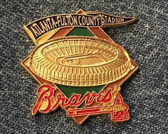 May include: A gold-colored Atlanta-Fulton County Stadium pin with a baseball stadium design. The pin features the word "Braves" in red script and a tomahawk. The pin is set against a denim background.