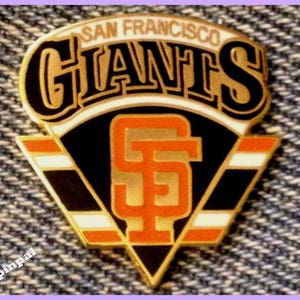 May include: A San Francisco Giants pin with the team's logo. The pin features the team name in black and orange, with a stylized "SF" logo in the center. The pin has a black, white, and orange color scheme.