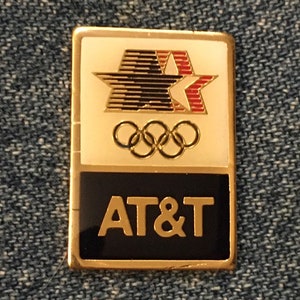 May include: A rectangular pin with a gold border. The top section is white with a black and red star design and Olympic rings. The bottom section is black with "AT&T" in gold lettering.
