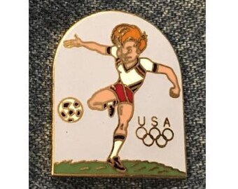 May include: A gold-colored pin featuring a cartoon soccer player kicking a ball. The player is wearing a white and red uniform with the word "USA" and the Olympic rings below.