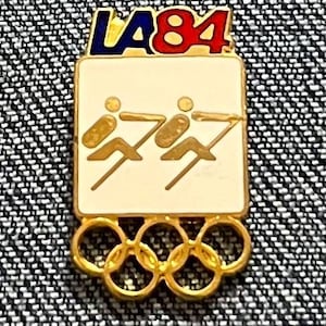 May include: Vintage LA 84 Olympic pin. The pin features the text "LA 84" in blue and red, above a cream-colored rectangle with two gold figures. Below the rectangle are the five gold Olympic rings. A collectible item.