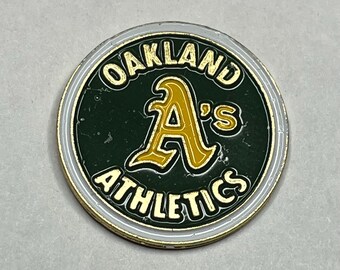 May include: A circular Oakland Athletics team logo pin. The pin features the team's logo in gold and white on a dark green background. The words "OAKLAND ATHLETICS" and "ATHLETICS" are printed around the logo.