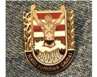 May include: Gold-colored pin with a swimmer diving into a wave. The pin features a red, white, and blue striped background with the text "XXIII Summer Games".