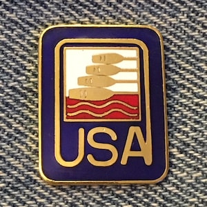 Puede incluir: Un pin de color dorado con fondo de esmalte azul. El pin presenta un gráfico blanco y rojo de tres remos en el agua con la palabra "USA" en letras doradas.