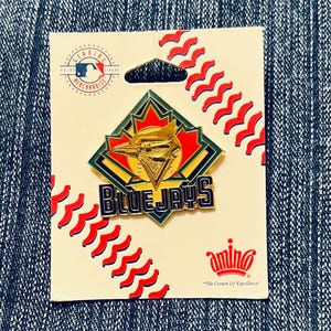 Puede incluir: Un pin de los Blue Jays en una tarjeta color crema. El pin presenta el logotipo del equipo en oro, azul, rojo y verde. La tarjeta tiene costuras de béisbol rojas y el logotipo de la MLB. Las palabras "Blue Jays" se muestran en el pin.