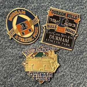 May include: Three gold-coloured pins featuring the Durham Bulls baseball team. The first pin is round and features the team's logo, the Durham Athletic Park, and the years 1939 and 1993. The second pin is rectangular and features the team's logo, the words "Durham Bulls", "1993", "Bulls", "vs Indians", and "Last Game-Sept 4th Durham Athletic Park". The third pin is a baseball diamond with the words "Durham Athletic Park" and "Durham Bulls".