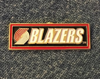 Puede incluir: Un pin de color dorado con un diseño rojo, blanco y negro. El pin presenta el logotipo de los Portland Trail Blazers y la palabra "BLAZERS" en letras blancas con un contorno dorado. El pin tiene un borde rojo y un borde dorado.