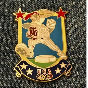 Peut inclure: Une épingle dorée représentant un lapin de dessin animé portant un uniforme de baseball et tenant un gant. Le lapin est debout sur une base de baseball avec une bannière qui dit "USA" et qui a cinq étoiles et les anneaux olympiques.