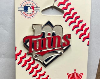 May include: A Twins pin on a cream-colored card with a red baseball stitch design. The pin features the word "Twins" in red lettering over a silver and navy blue background. A red pin is attached to the card.