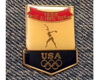 May include: Gold and red pin with a stylized figure of a woman throwing a javelin. The pin reads "1988 US Olympic Team" and "USA" with the Olympic rings below.