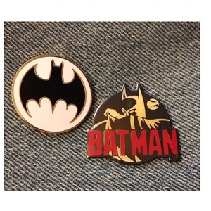 Op de afbeelding: Twee Batman-speldjes. E&eacute;n is een zwart-wit silhouet van het Batman-logo. De andere is een zwart-gouden silhouet van Batman met het woord "BATMAN" in rode letters.